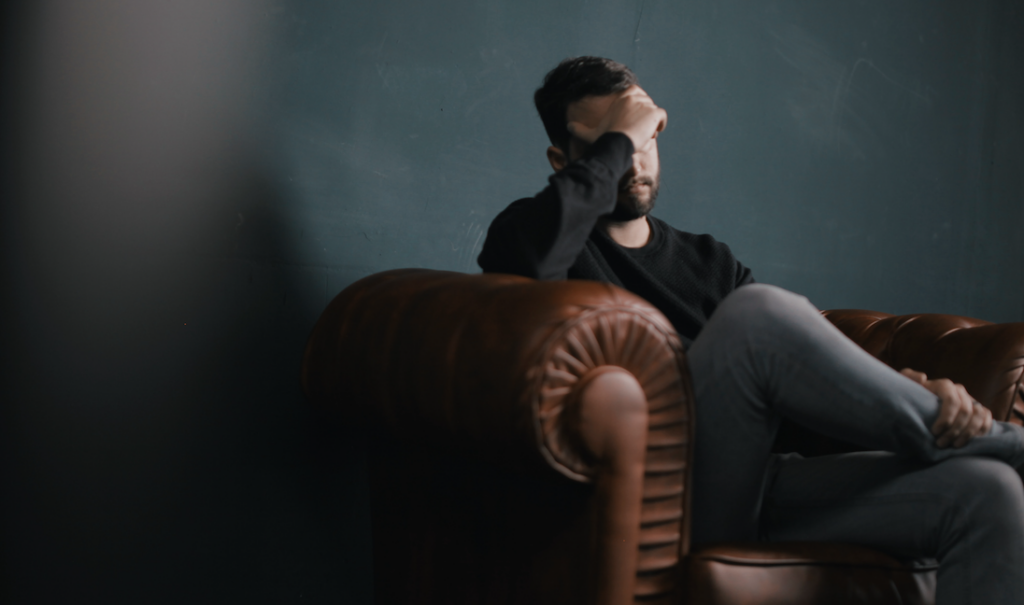 man sitting on sofa with hand over eyes - appears stressed and dealing with burnout