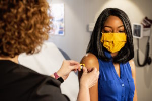 covid-19 vaccine - AINsight article - Sheryl Barden - black woman receiving vaccine from white nurse